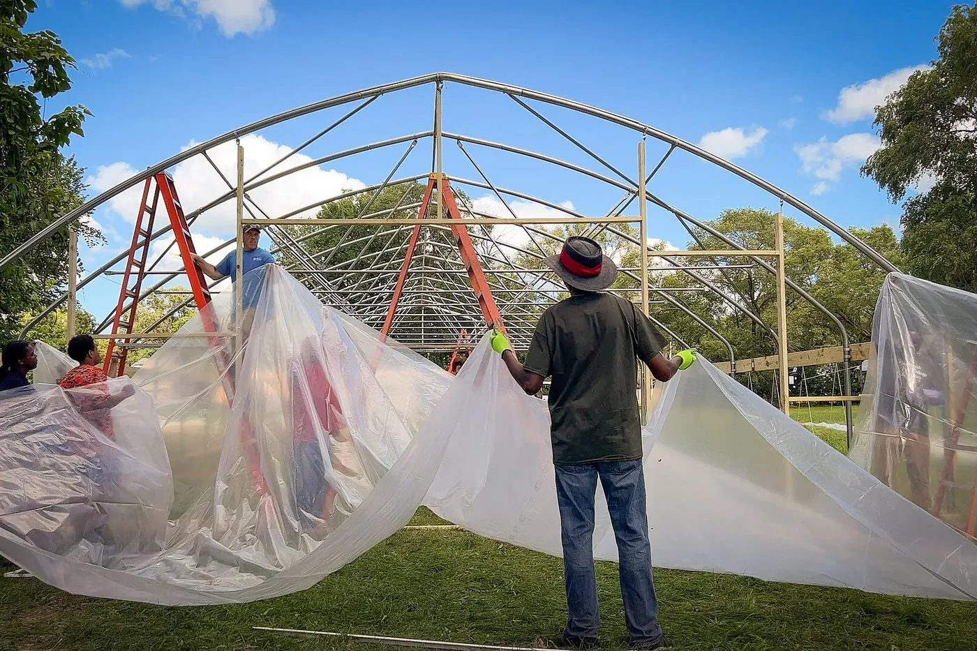 A man in a hat stretching plastic sheeting across an arched frame, his back to the camera.