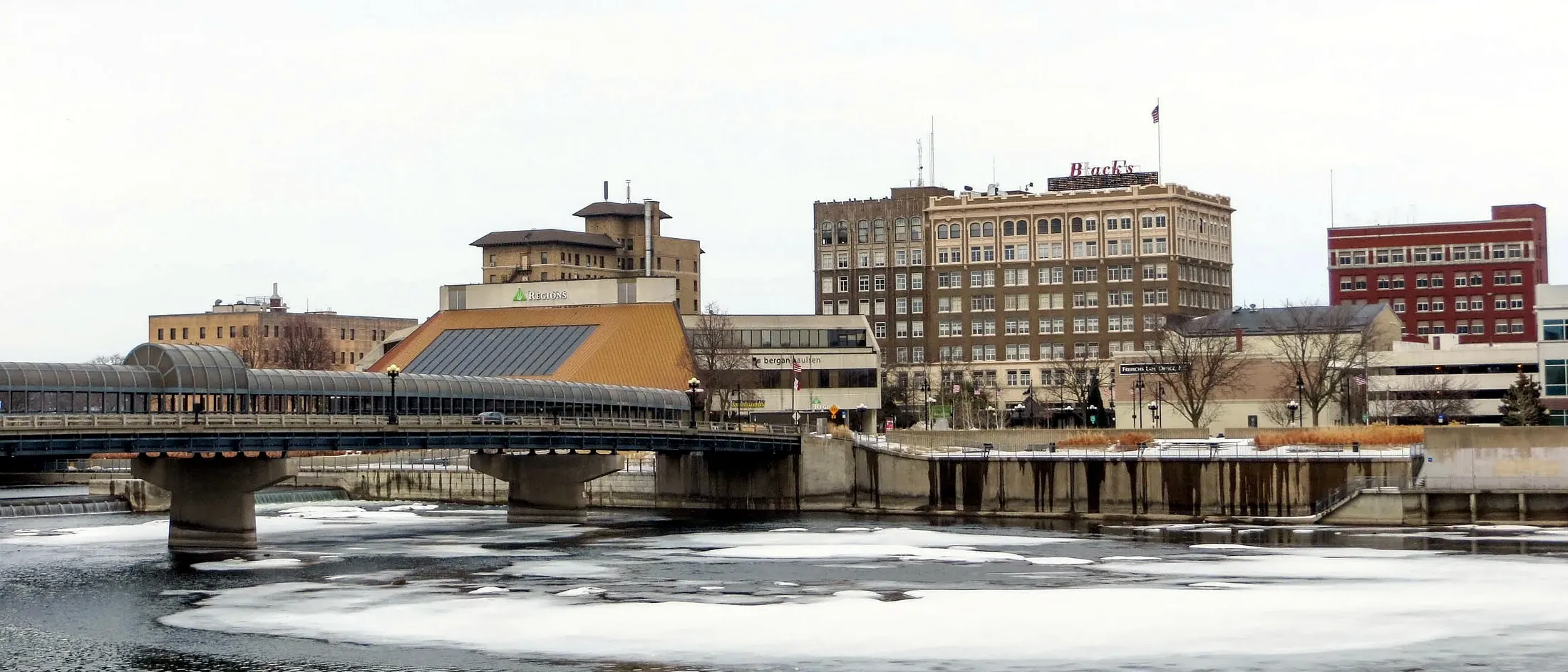 Downtown Waterloo seen across the Cedar River in winter, with a bridge spanning the water.