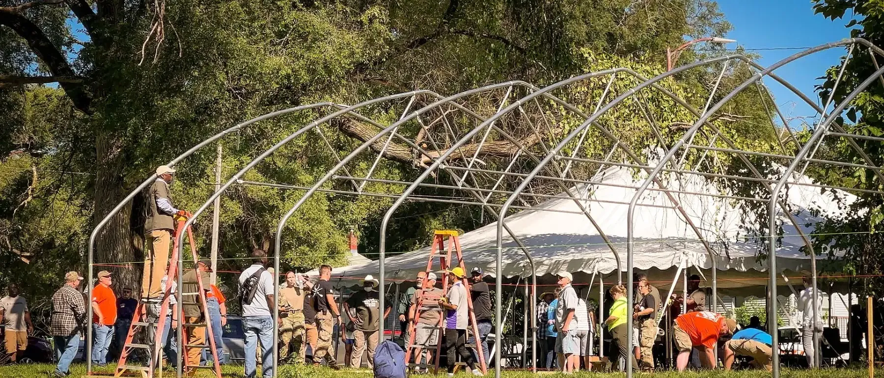 Rows of steel arches rising over a grassy lot as a crowd of volunteers builds a greenhouse.